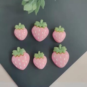 May include: Six pink strawberry-shaped embellishments with green tops are arranged on a dark gray surface. The strawberries have white speckles and are of varying sizes. A few green leaves are visible in the background.