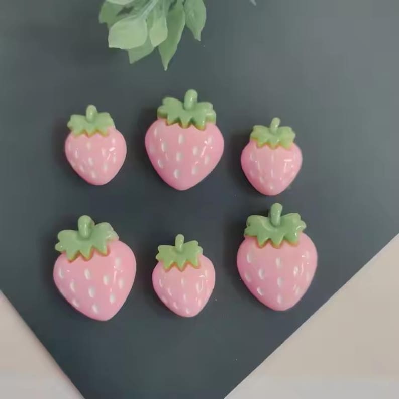 May include: Six pink strawberry-shaped embellishments with green tops are arranged on a dark gray surface. The strawberries have white speckles and are of varying sizes. A few green leaves are visible in the background.