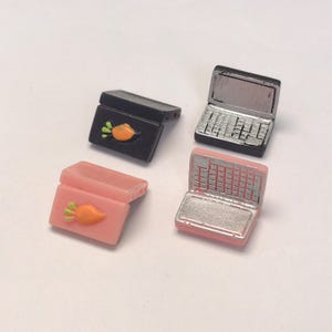 May include: Four miniature laptop and case charms. Two are black, one with a carrot design. The other two are pink, one with a carrot design. The laptops have silver keyboards and screens.
