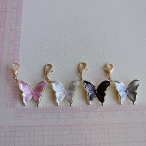 May include: Five butterfly charms in shades of pink, white, black, and gray, each with a gold-colored clasp. The charms are arranged in a row, with a ruler in the background, showing measurements in centimeters.