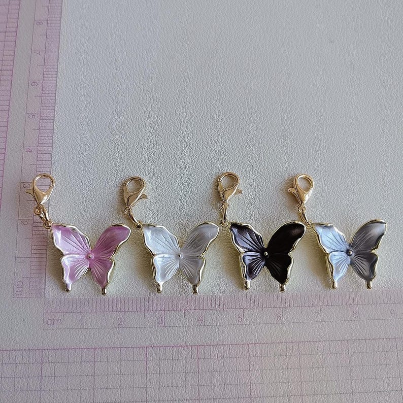 May include: Five butterfly charms in shades of pink, white, black, and gray, each with a gold-colored clasp. The charms are arranged in a row, with a ruler in the background, showing measurements in centimeters.
