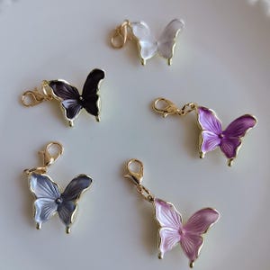 May include: Five butterfly charms in various colors: white, black, purple, gray, and pink. Each charm has a gold-colored border and a lobster clasp for attachment. The butterfly wings have a translucent, layered appearance.
