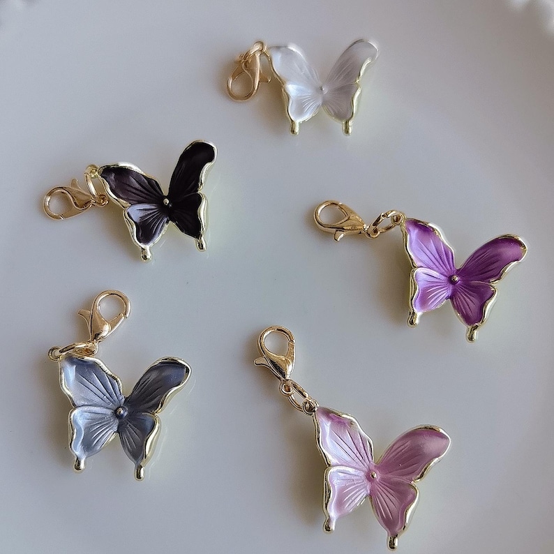 May include: Five butterfly charms in various colors: white, black, purple, gray, and pink. Each charm has a gold-colored border and a lobster clasp for attachment. The butterfly wings have a translucent, layered appearance.