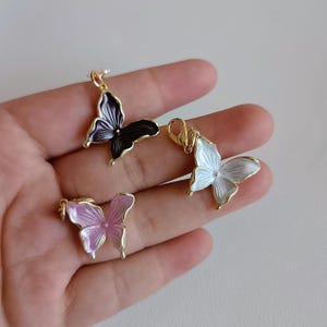 May include: Three butterfly pendants in gold, black, white, and pink. The butterfly charms have a gold bail and are displayed against a neutral background. The pendants are likely for jewelry making or crafting.