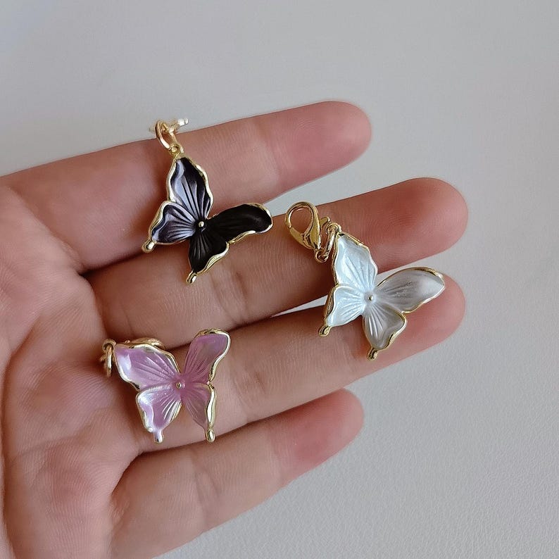 May include: Three butterfly pendants in gold, black, white, and pink. The butterfly charms have a gold bail and are displayed against a neutral background. The pendants are likely for jewelry making or crafting.