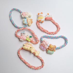 May include: Five colorful hair ties with braided bands. Each tie features a different cartoon character, including a bear, bunny, and duck. The characters are in pastel colors, and the bands are pink, blue, and yellow.