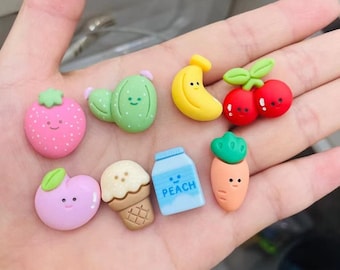 May include: A set of nine colorful, cartoon-style food charms, including a strawberry, a cactus, a banana, a cherry, a carrot, a peach-flavored milk carton, a peach, an ice cream cone, and a strawberry.