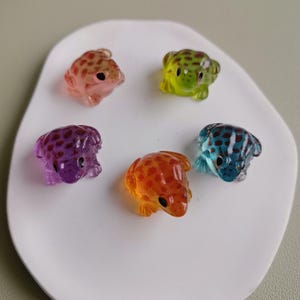 Può includere: Una collezione di sei piccole figurine a forma di rana traslucide in vari colori, tra cui rosa, verde, viola, arancione e blu. Ogni rana ha un motivo a pois e occhi neri, disposte su una superficie bianca di forma organica.