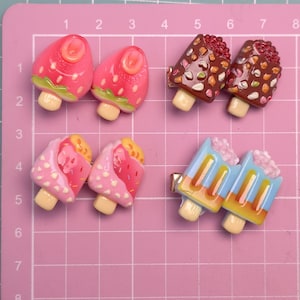 Pode incluir: Seis grampos de cabelo decorativos em forma de sobremesas em miniatura. Os grampos apresentam desenhos de morangos, chocolate e picolés. Os grampos estão em um fundo rosa quadriculado.