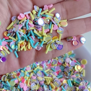 May include: A close-up of a hand holding a mix of colorful polymer clay sprinkles. The sprinkles include pastel yellow sticks, light blue cylinders, pink hearts, and round, iridescent pieces. There are also small, decorated egg-shaped pieces.