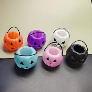 Könnte beinhalten: Eine Sammlung kleiner, farbenfroher Kürbis-Eimer aus Kunststoff mit schwarzen Griffen. Die Eimer sind in verschiedenen Farben erhältlich, darunter Orange, Lila, Weiß, Blau, Rosa und Schwarz, jeweils mit einem geschnitzten Kürbisgesicht. Ideal für Halloween.