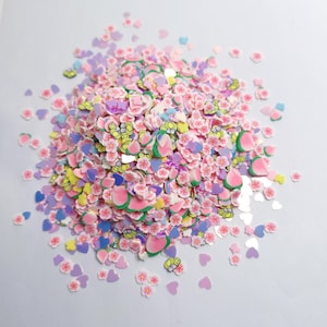 Puede incluir: Una colección de pequeñas rodajas decorativas planas de arcilla polimérica en varias formas, incluyendo flores de cerezo rosas, mitades de melocotón, abejas amarillas y corazones morados. Estos coloridos adornos artesanales están esparcidos.