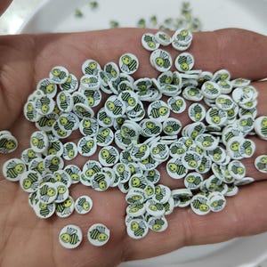 May include: A scattering of small, white, circular embellishments with a yellow and black bee design. The bees have smiling faces. These decorative pieces are suitable for crafts and DIY projects.