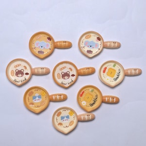 May include: Nine small, decorative frying pans with cartoon animal designs. Each pan has a different design, including a bear, koala, and cat, with the words "Bear Cook" and "Breakfast" on some. The pans are yellow and white with brown handles.