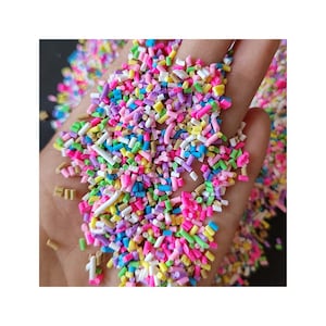 May include: A close-up of a hand filled with colourful sprinkles. The sprinkles are small, rectangular shapes in shades of pink, blue, yellow, green, and purple. Ideal for decorating cakes and other treats. The image is brightly lit, showing the vibrant colours.