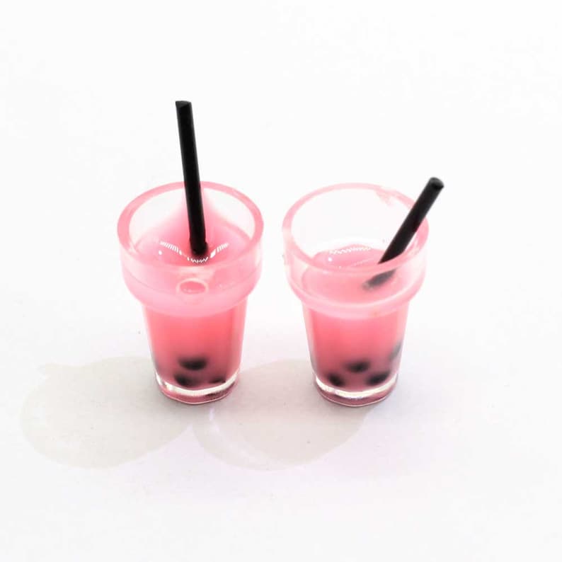 May include: Two miniature pink bubble tea drinks with black straws in clear plastic cups. The drinks have black tapioca pearls at the bottom.