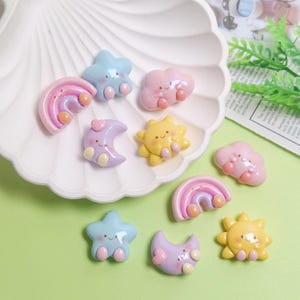 Puede incluir: Una colección de colgantes de resina de colores pastel caprichosos con forma de arcoíris, estrella, nube, luna y sol. Cada colgante tiene una cara sonriente y pies pequeños. Los colgantes se muestran en un plato blanco con volantes.