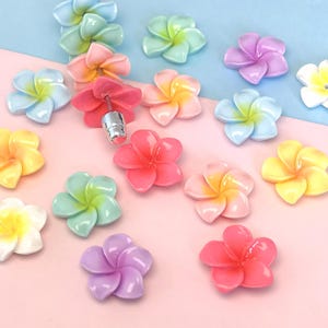 Może przedstawiać: Kolekcja kolorowych ozdób kwiatowych plumerii z żywicy w różnych odcieniach różu, żółci, niebieskiego, zielonego i fioletowego. Niektóre kwiaty mają srebrny metalowy słupek. Kwiaty ułożone są na różowo-niebieskim tle.