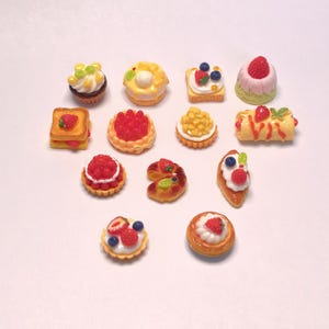 May include: A selection of miniature pastries, including tarts, cakes, and sandwiches, with various fruit toppings. The desserts feature strawberries, blueberries, and other colourful elements. The arrangement is on a white surface.