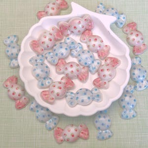 May include: A collection of candy-shaped decorations in pink and blue, with a strawberry pattern, arranged on and around a white seashell-shaped dish. The candies have a glossy finish and are scattered on a light green surface.
