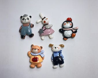 May include: A collection of five ceramic figurines featuring animal characters. The figurines include a panda bear, a rabbit, a penguin, a teddy bear, and a dog. Each figure is wearing clothing and has a unique design, with a variety of colors and patterns.