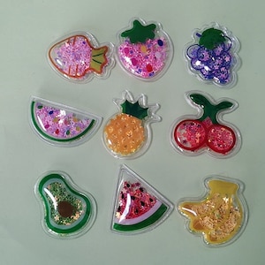 Könnte beinhalten: Neun bunte, fruchtig geformte Harzsticker mit Glitzerakzenten. Die Sticker zeigen einen Ananas, eine Wassermelone, eine Erdbeere, eine Kirsche, eine Avocado, Trauben und eine Karotte.