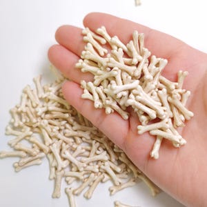 May include: A collection of small, off-white bone-shaped decorative pieces. Some of the bones are held in a hand, while others are scattered on a white surface. The bones have a slightly aged appearance, with subtle brown shading.