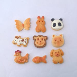 May include: A collection of nine animal-shaped resin embellishments. The assortment includes a butterfly, rabbit, panda, cow, monkey, cat, chicken, fish, and dog. Each piece features a detailed, cartoon-like design with varying shades of orange, brown, and white.