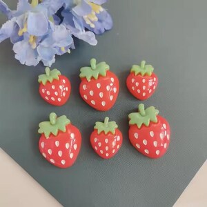 May include: Six red strawberry-shaped embellishments with green tops and white seed details. The strawberries are arranged on a gray surface, with blue flowers in the background. These decorative pieces are ideal for crafts.