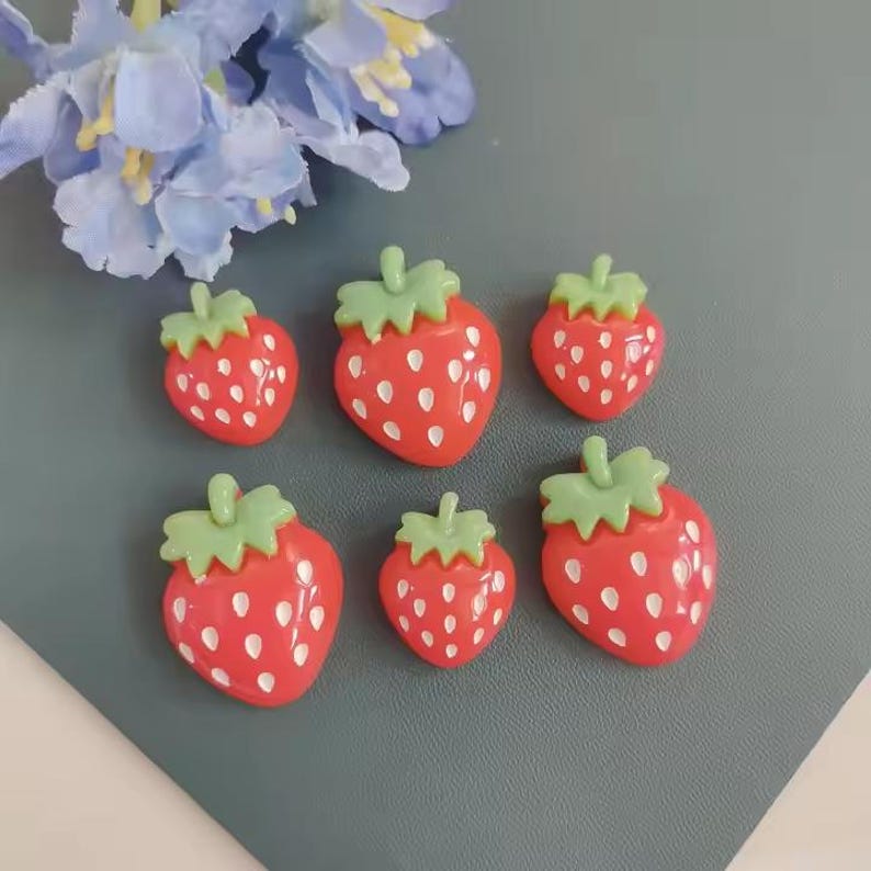 May include: Six red strawberry-shaped embellishments with green tops and white seed details. The strawberries are arranged on a gray surface, with blue flowers in the background. These decorative pieces are ideal for crafts.