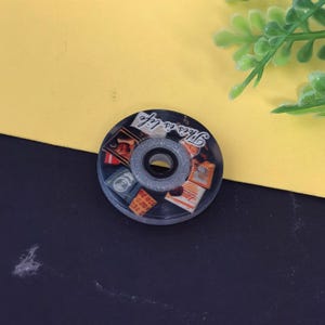 May include: A round, black and clear roller skate wheel with a central bearing. The wheel features a collage of vintage-style images and the phrase "This is life" in white script.