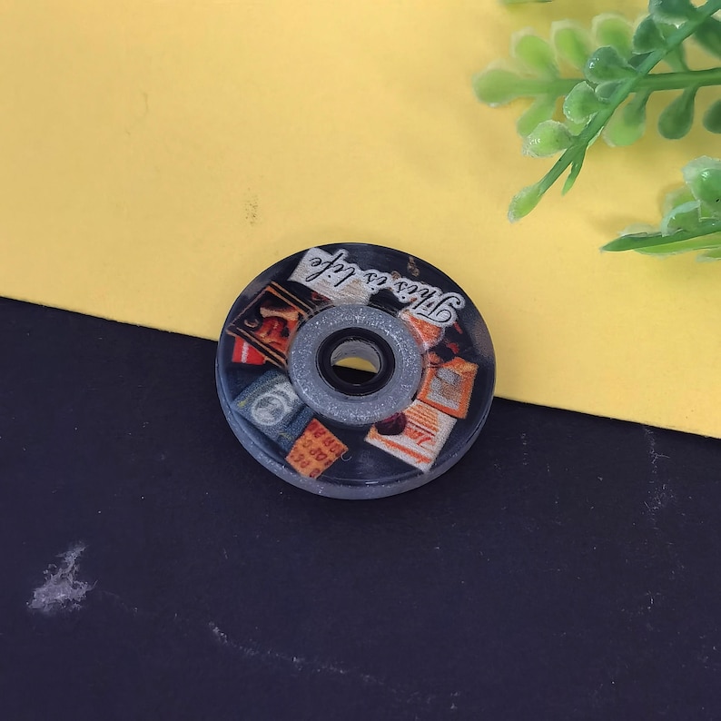 May include: A round, black and clear roller skate wheel with a central bearing. The wheel features a collage of vintage-style images and the phrase "This is life" in white script.
