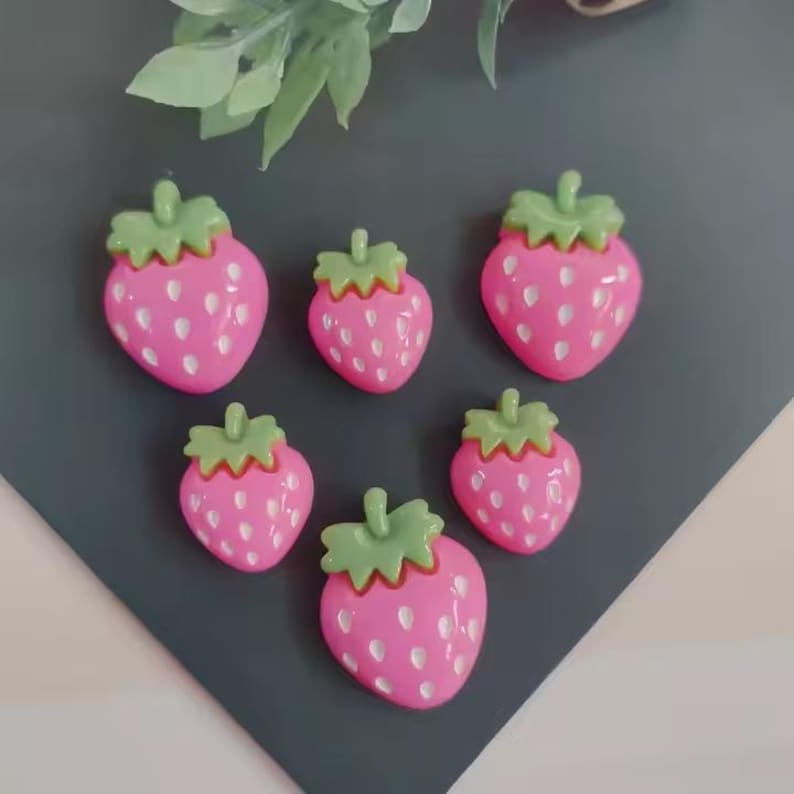 May include: Six pink strawberry-shaped embellishments with green tops and white seed details. The strawberries are arranged on a dark gray and white surface, with a blurred green leafy background.
