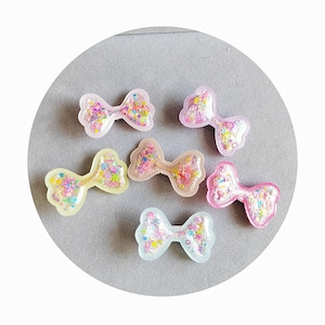 May include: Six colorful resin bows with glitter embedded inside. The bows are in various colors, including pink, blue, yellow, and green.