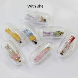 Op de afbeelding: Acht transparante plastic capsules met miniatuurfiguren erin. De figuren dragen kleding in verschillende kleuren. De tekst "With shell" staat boven de capsules.