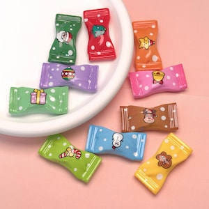 May include: A collection of colorful, miniature candy-shaped ornaments. Each ornament features a different festive design, including a Santa Claus, a Christmas tree, and a reindeer. The ornaments are arranged on a white plate and a pink surface.