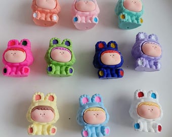 May include: A collection of colorful, small figurines shaped like a person wearing a hooded animal costume. The figurines are in various colors, including pink, green, blue, and yellow. The faces are pale with small dots for eyes and a small mouth.