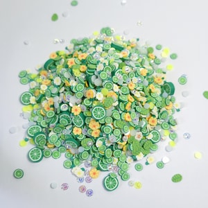 Puede incluir: Una colección de pequeñas rodajas de arcilla polimérica con forma de gajos de lima verde, flores amarillas y hojas verdes, mezcladas con diminutas lentejuelas circulares blancas iridiscentes. Las piezas están esparcidas en un montón.