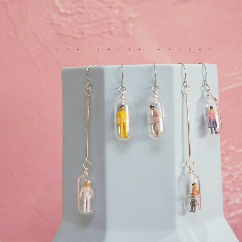 Op de afbeelding: Vier paar zilveren oorhangers met heldere glazen capsules die miniatuurfiguren bevatten. De figuren dragen kleding in verschillende kleuren. De oorhangers worden gepresenteerd op een lichtblauw oppervlak.