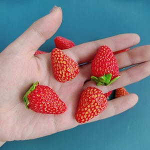 Könnte beinhalten: Mehrere rote, erdbeerförmige Objekte mit grünen Blättern werden gezeigt. Die Erdbeeren haben eine strukturierte Oberfläche mit gelben Samen. Die Objekte werden in einer offenen Hand vor blauem Hintergrund gehalten.