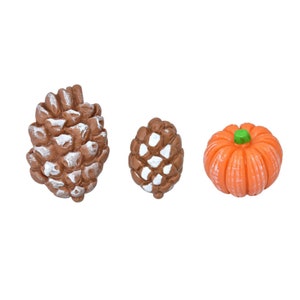 Puede incluir: Tres decoraciones de otoño en miniatura: una piña marrón, una piña blanca más pequeña y una calabaza naranja.