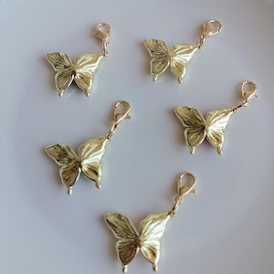 May include: Five gold-colored butterfly charms with lobster clasp attachments. The charms have detailed wing patterns and are arranged on a white surface. These decorative charms are suitable for jewelry making or craft projects.