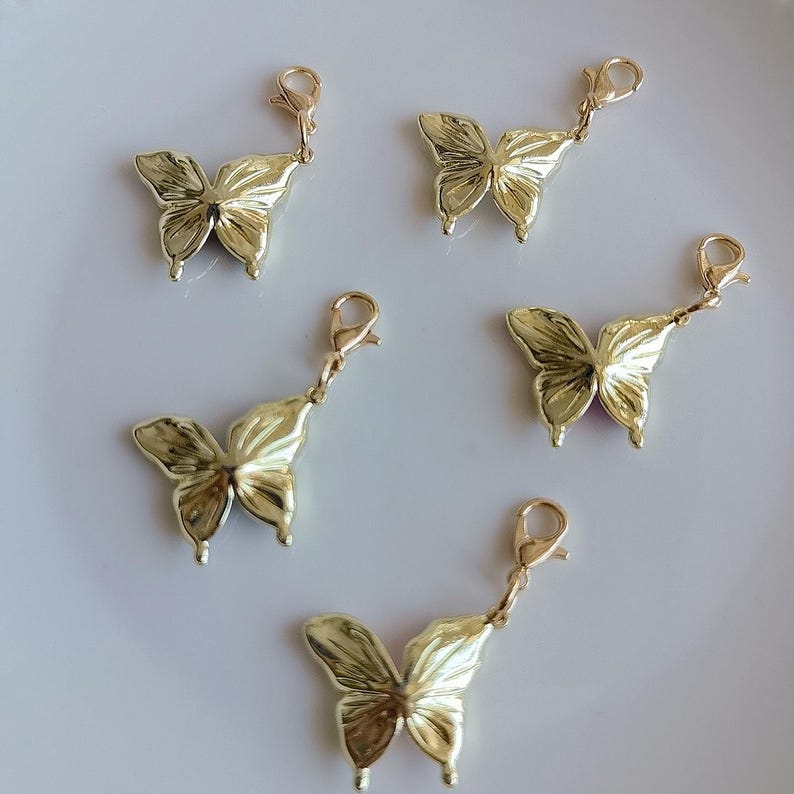 May include: Five gold-colored butterfly charms with lobster clasp attachments. The charms have detailed wing patterns and are arranged on a white surface. These decorative charms are suitable for jewelry making or craft projects.