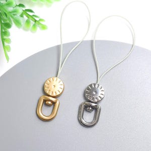 Peut inclure: Deux attaches de rideaux. L'une est dorée, l'autre argentée. Chacune a un dessus circulaire avec un motif en forme d'éclat de soleil et un bas en forme de U. Un cordon blanc relie le haut au bas. Les attaches sont sur une surface grise.