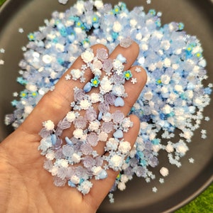 May include: A close-up of a hand holding a variety of blue and white resin sprinkles. The sprinkles are shaped like flowers, hearts, and stars. The sprinkles are scattered on a black surface.