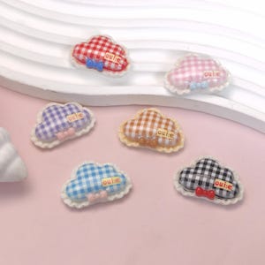 May include: A collection of cloud-shaped embellishments with gingham patterns in various colors, including red, pink, blue, purple, brown, and black. Each cloud has a small bow and the text "oui:e". The clouds are trimmed with a white, scalloped edge.