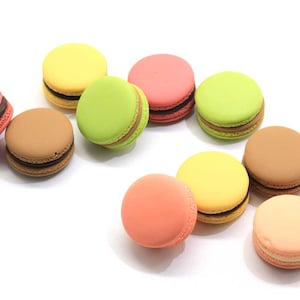 May include: Assortment of colorful macarons, including yellow, green, pink, brown, and peach. The macarons are arranged on a white surface.