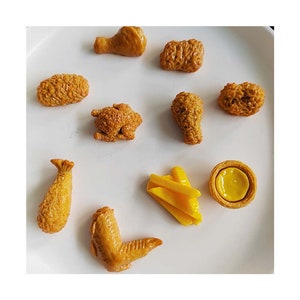 Peut inclure: Une collection de morceaux de poulet frit miniatures et une petite tarte sur une assiette blanche. Les morceaux de poulet sont de formes et de tailles variées, notamment des cuisses, des ailes et des pépites. La tarte est une petite pâtisserie ronde avec une croûte dorée et une garniture jaune.
