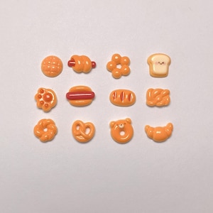 May include: A collection of miniature orange-colored food-shaped charms. The charms include a waffle, pretzel, flower-shaped pastry, bread slice, hot dog, baguette, and a bear-shaped donut. These charms are likely for crafting or jewelry making.