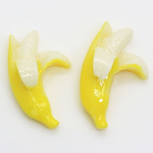 Op de afbeelding: Twee gele bananenvormige bedels met witte details. De bedels zijn gemaakt van een glad, glanzend materiaal en perfect om elke knutselproject een leuke en speelse touch te geven.
